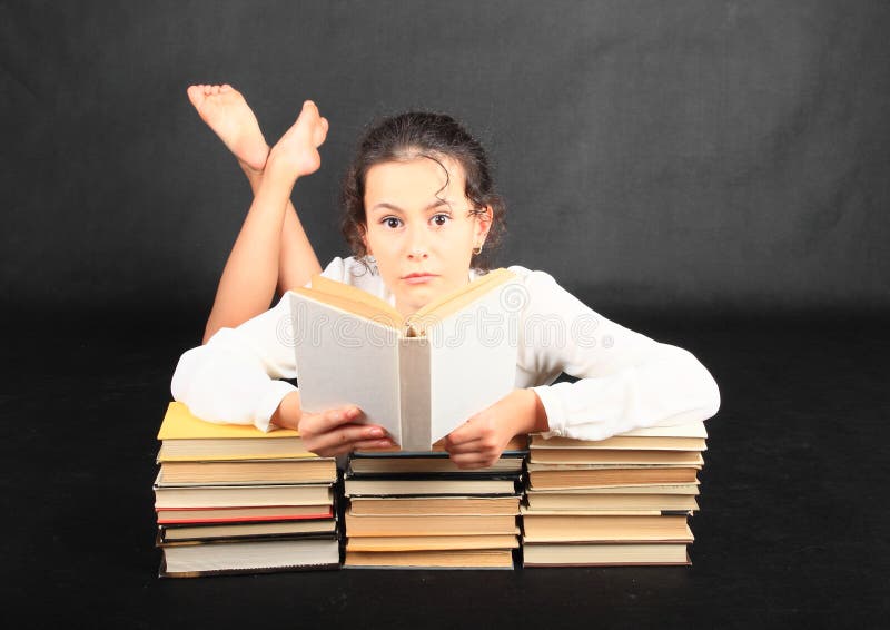 Pretty teenage girl - barefoot schoolkid reading a book while leaning on stacks of old books on black background. Entertainment and education concept. Pretty teenage girl - barefoot schoolkid reading a book while leaning on stacks of old books on black background. Entertainment and education concept.