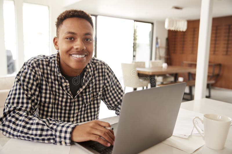 Adolescente masculino afroamericano que usa un ordenador portátil en casa que sonríe a la cámara, cierre para arriba