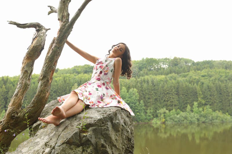 Pretty teenage girl - smiling barefoot schoolkid with brunette hair dressed in nice light summer dress sitting on rock above river. Outdoor portrait photography concept. Pretty teenage girl - smiling barefoot schoolkid with brunette hair dressed in nice light summer dress sitting on rock above river. Outdoor portrait photography concept.