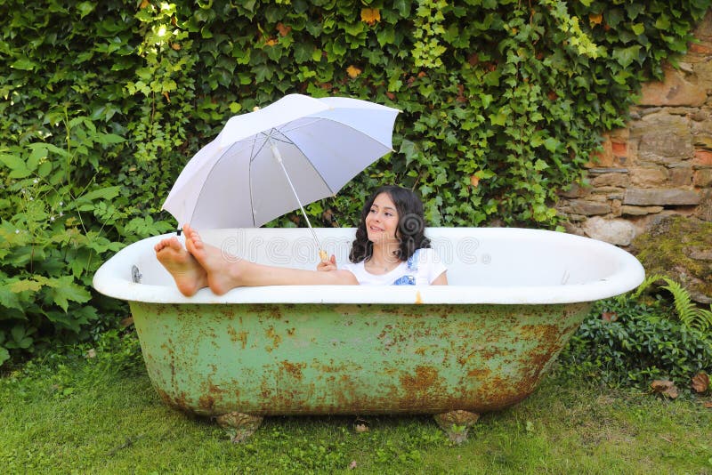 Smiling teenage girl - barefoot schoolkid lying in an old bathtub with bare feet out holding white umbrella. Outdoor with wall overgrown with ivy behind. Retro home related portrait concept. Smiling teenage girl - barefoot schoolkid lying in an old bathtub with bare feet out holding white umbrella. Outdoor with wall overgrown with ivy behind. Retro home related portrait concept.