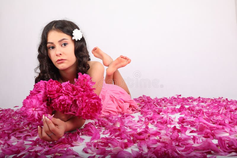 Melancholic teenage girl - schoolkid with white flower in brunette hair dressed in pink dress holding bouquet of peonies lying on petals of pink peonies. Pink innocence concept. Melancholic teenage girl - schoolkid with white flower in brunette hair dressed in pink dress holding bouquet of peonies lying on petals of pink peonies. Pink innocence concept.