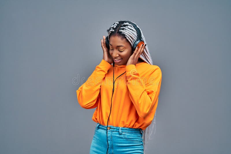Adorabile donna americana in camicia gialla che canta e si diverte mentre ascolta della musica usando cuffie isolate in grigio fotografia stock