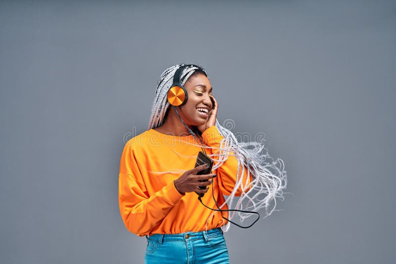 Adorabile donna americana con camicia gialla che danza e si diverte mentre ascolta della musica usando cuffie isolate in grigio fotografia stock