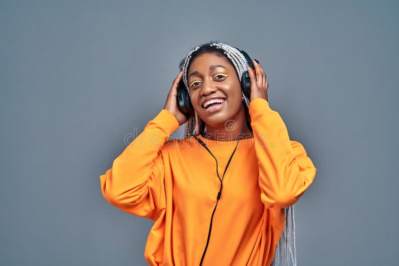 Adorabile per una donna americana sorridente con camicia gialla che ascolta musica usando cuffie wireless isolate su fondo grigio immagine stock libera da diritti