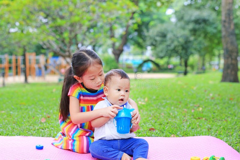 Adorable Asian sister sitting on pink mattress mat take care her little brother to drinking water from Baby sippy cup with straw