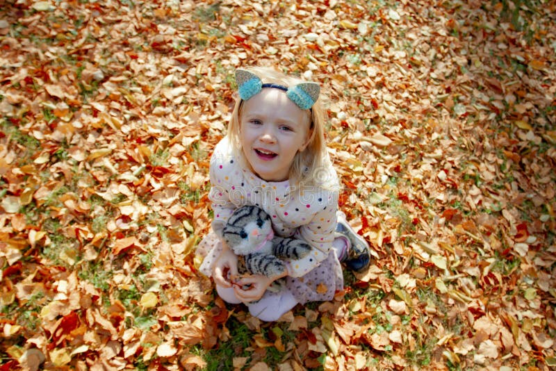 Adorable blond girl sitting on yellow fallen leaves looking up on a beautiful autumn day.