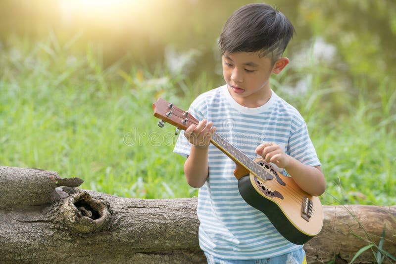 Adorable boy with guitar sitting on the grass on sunset, Musical concept with little boy playing ukulele at sunny park