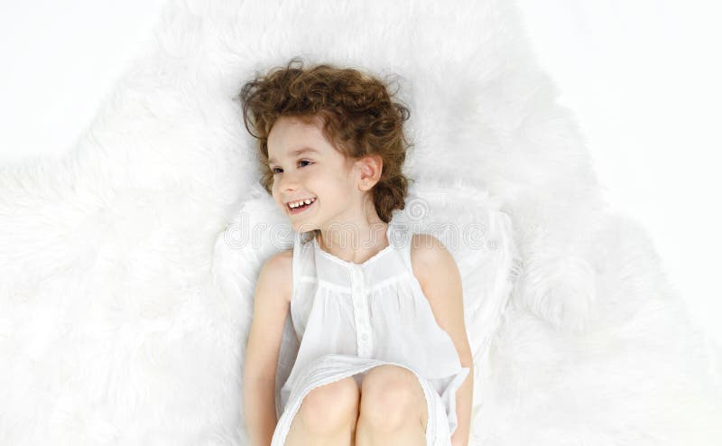 Adorable curly little girl lying down on fur, looking at a side, in white dress and wings, isolated on white background. stock photography Adorable curly little girl lying down on fur, looking at a side, in white dress and wings, isolated on white background. stock photography