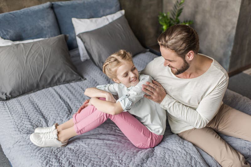 Happy adorable daughter and father sitting on bed at home.