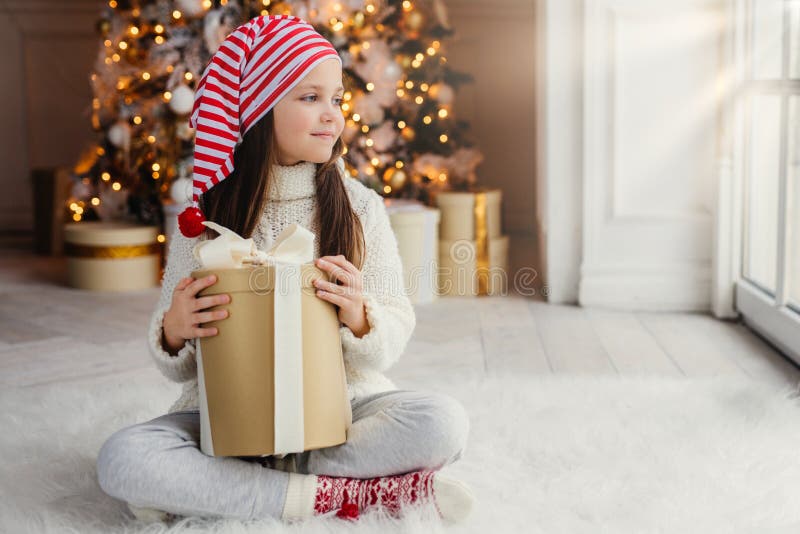 Adorable female child wears santa claus hat, warm clothes, hold wrapped gift box, sits against decorated New Year tree. Happy pret royalty free stock images Adorable female child wears santa claus hat, warm clothes, hold wrapped gift box, sits against decorated New Year tree. Happy pret royalty free stock images