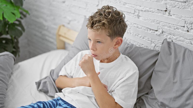 Adorable garçon blond assis sur son lit dans son pyjama toussant tôt le matin. enfant malade se reposant dans la chambre à coucher
