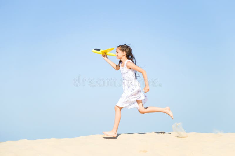 Adorable happy little girl wearing a white dress running on the sandy beach by the sea and playing with the y