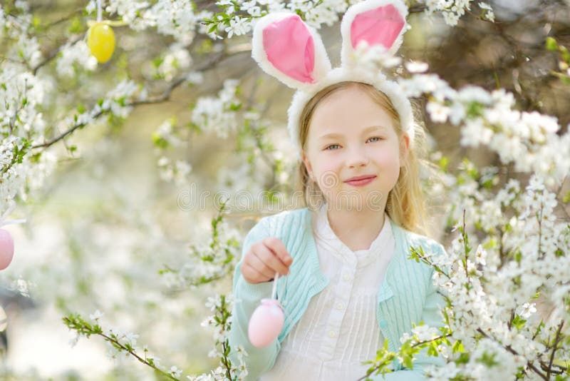 Adorable little girl wearing bunny ears in blooming cherry garden on beautiful spring day. Kid hanging Easter eggs on blossoming