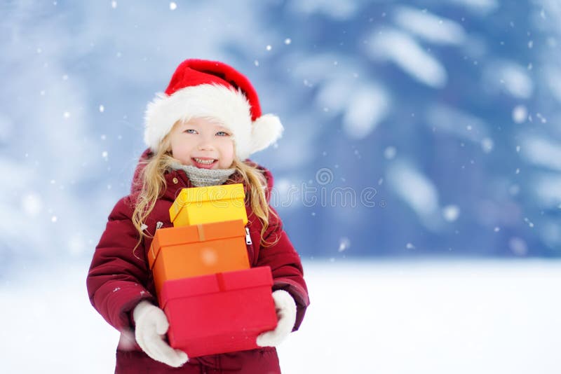 Adorable little girl wearing Santa hat holding a pile of Christmas gifts on beautiful winter day