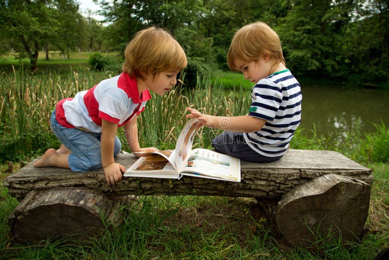 Adorable Little Twin Brothers Sitting on a Wooden Bench and Looking at Interesting Pictures in the Book Near the Beautiful Lake