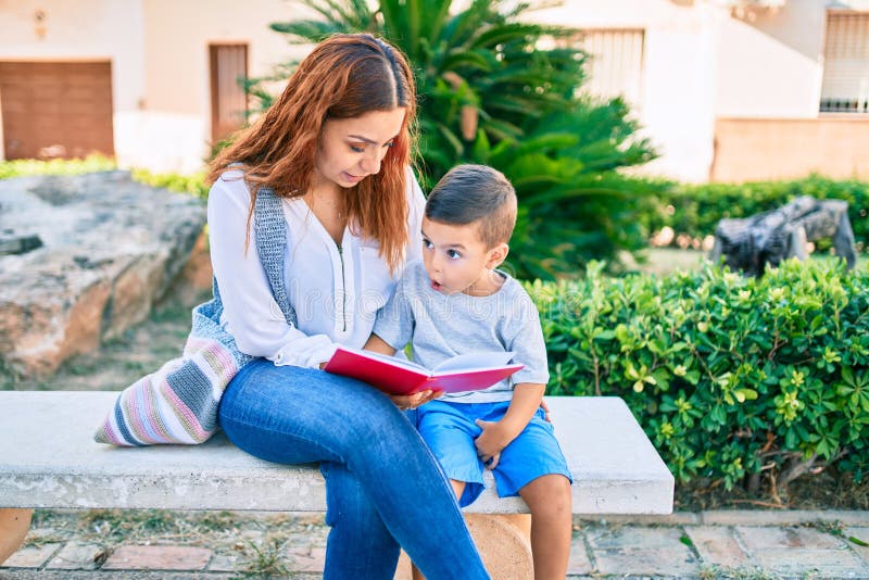 Adorable madre latina e hijo sentados en el banco y leyendo el libro en el parque imagen de archivo