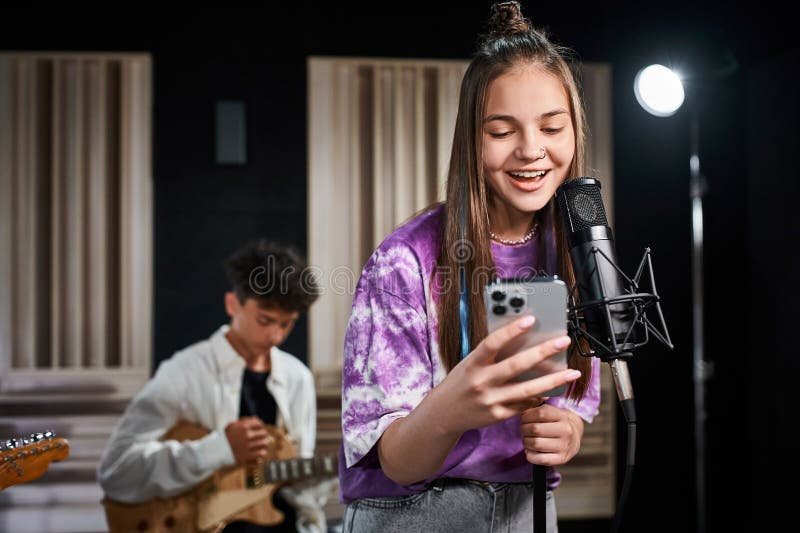 jolly adorable teenage girl singing and looking at smartphone next to her blurred guitarist, stock photo. jolly adorable teenage girl singing and looking at smartphone next to her blurred guitarist, stock photo