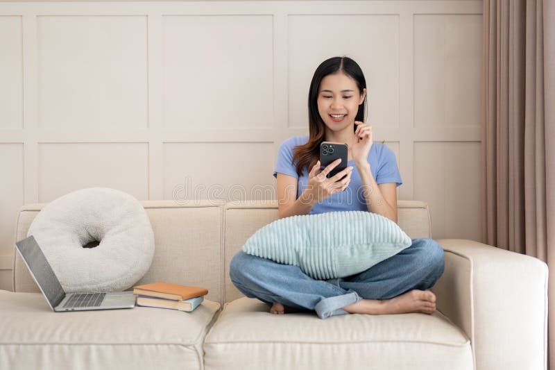 Adorable mujer asiática sentada en el sofá mientras usa un smartphone en la sala de estar fotos de archivo