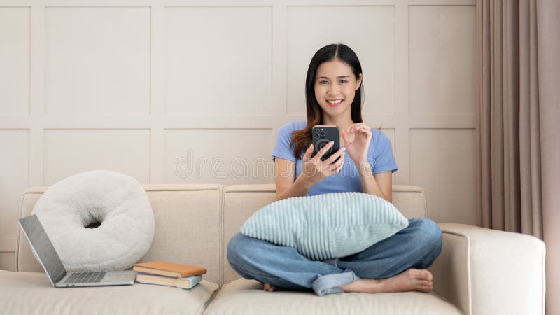 Adorable mujer asiática sentada en el sofá mientras usa un smartphone en la sala de estar foto de archivo libre de regalías