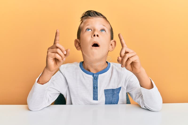 Adorable caucasian kid wearing casual clothes sitting on the table amazed and surprised looking up and pointing with fingers and raised arms. Adorable caucasian kid wearing casual clothes sitting on the table amazed and surprised looking up and pointing with fingers and raised arms