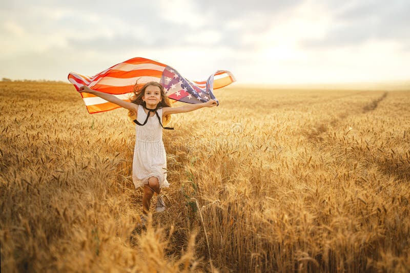 Adorable patriotic girl in white dress wearing an American flag while running in a beautiful wheat field.