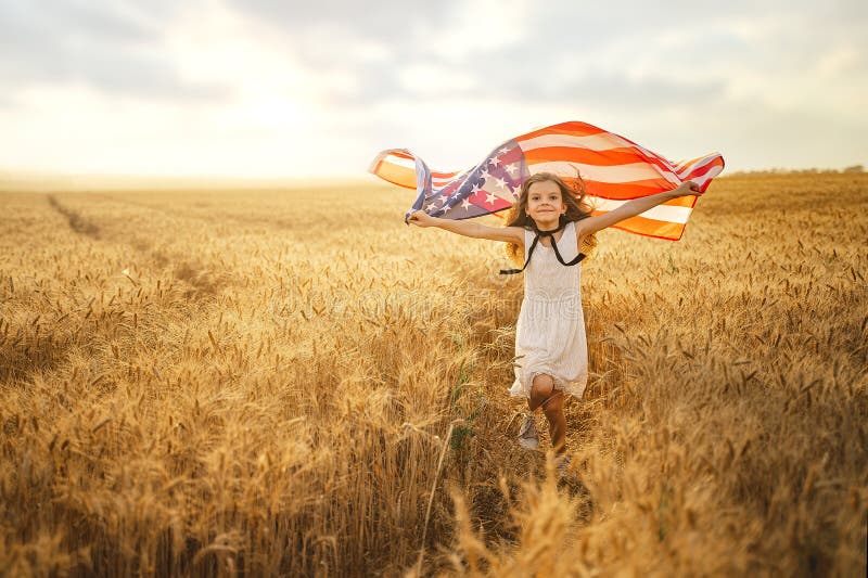 Adorable patriotic girl in white dress wearing an American flag while running in a beautiful wheat field