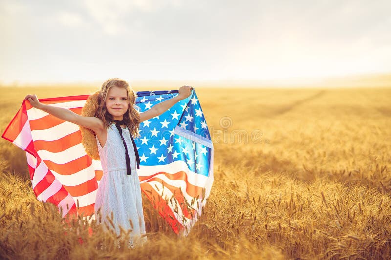 Adorable patriotic girl in white dress wearing an American flag while running in a beautiful wheat field.