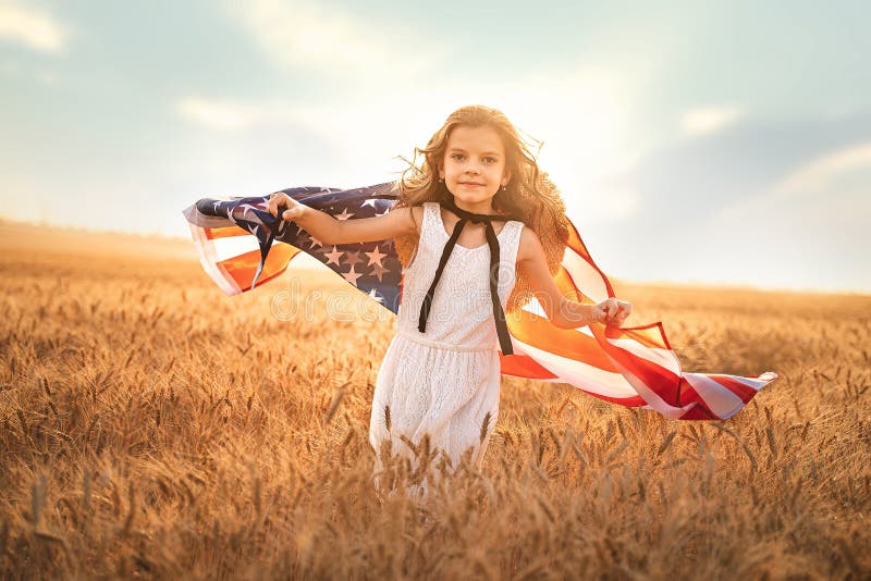 Adorable patriotic girl in white dress wearing an American flag while running in a beautiful wheat field