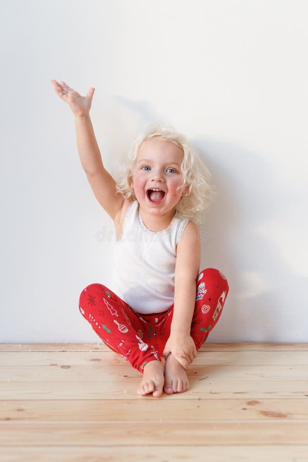 Adorable small female child wears pyjamas, sits on wooden floor raises hands as happy to see affectionate parents stock photo Adorable small female child wears pyjamas, sits on wooden floor raises hands as happy to see affectionate parents stock photo