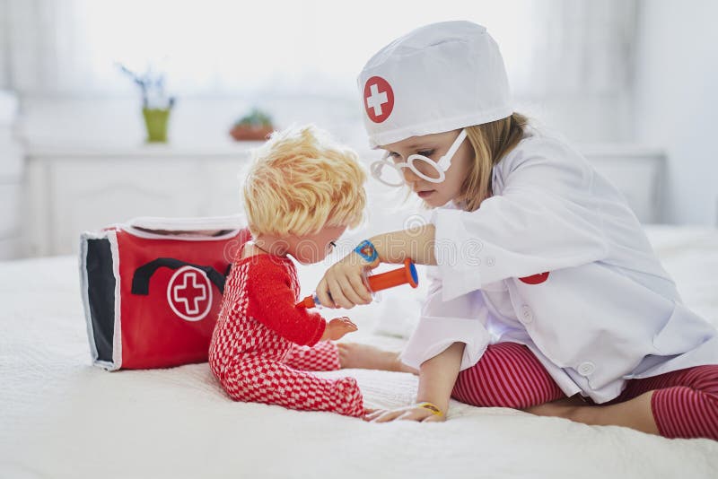 Adorable toddler girl in white coat playing doctor and giving medical care to her doll