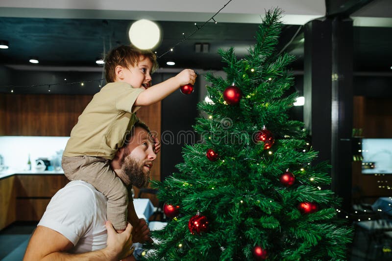 Adorable toddler son sitting on dad& x27;s shoulders decorating a christmas tree