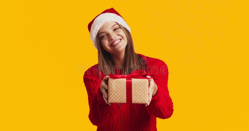 Adorable young lady in Santa hat and red sweater offering gift box to camera and smiling, orange studio background