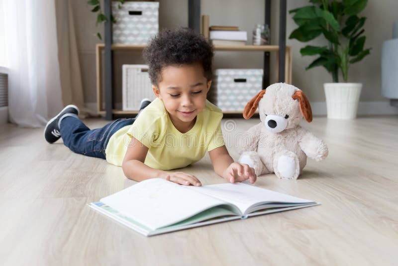 Adorable toddler mixed race boy reading book at home or in kindergarten, beautiful preschooler child with toy lying on warm floor, playing, having fun, kid and study, education concept. Adorable toddler mixed race boy reading book at home or in kindergarten, beautiful preschooler child with toy lying on warm floor, playing, having fun, kid and study, education concept