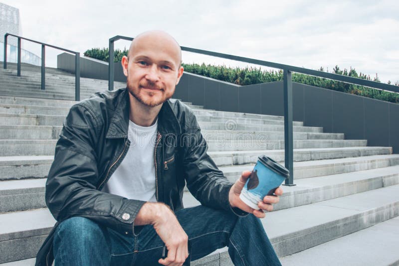 Adult bald bearded smiling man drinking coffee from paper cup and sitting in stair at city street royalty free stock images Adult bald bearded smiling man drinking coffee from paper cup and sitting in stair at city street royalty free stock images