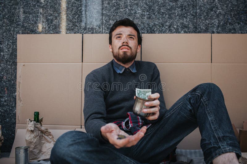 Adult beggar is sitting on cardboard and begging money. He is looking up. Man has some coins in one hand and a cup with