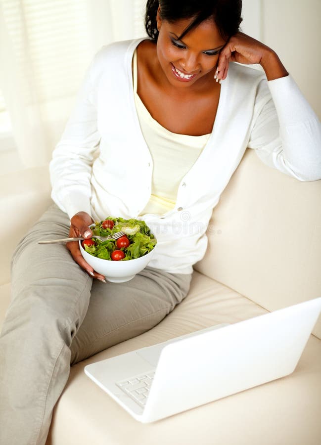 Top view portrait of an adult black woman eating healthy green salad while is sitting on sofa in front of her laptop