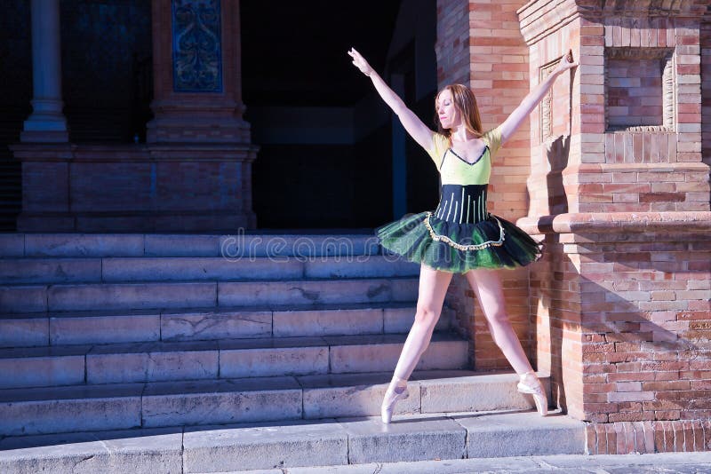 Adult female Hispanic classical ballet dancer in green and black tutu with coins, performing figures beside steps next to a brick
