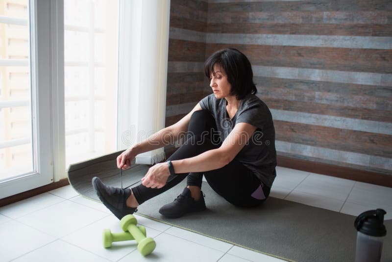 Adult fit slim woman has workout at home. Serious hardworking mature model sit on yoga mat to tie shoe laces. Getting