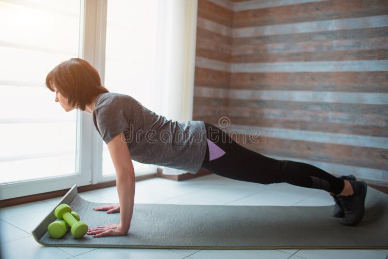 Adult fit slim woman has workout at home. Side view of strong well-built senior model stand in full plank position