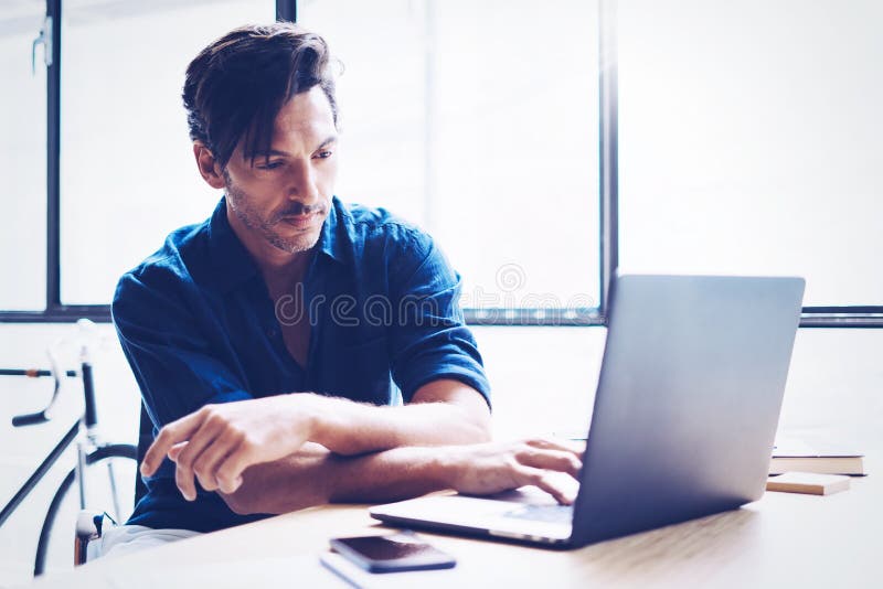 Adult handsome businessman working contemporary notebook while sitting at the wooden table at sunny office.Blurred royalty free stock image Adult handsome businessman working contemporary notebook while sitting at the wooden table at sunny office.Blurred royalty free stock image