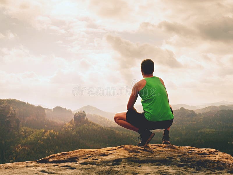 Adult hiker in black shorts and green singlet sit on mountain edge. Man enjoying view and looking to misty horizon