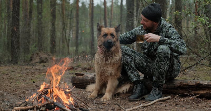 Adult Man is relaxing at the fire with his dog in the nature. Alone traveler is resting with his German shepherd by the forest