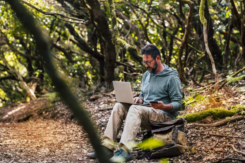 Adult man work with computer laptop and roaming phone connection in the middle of the green wild forest - concept of digital nomad