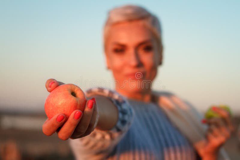 Adult middle-aged blonde woman in a sweater eating an apple and holding out another one to the camera, selective focus royalty free stock photo Adult middle-aged blonde woman in a sweater eating an apple and holding out another one to the camera, selective focus royalty free stock photo