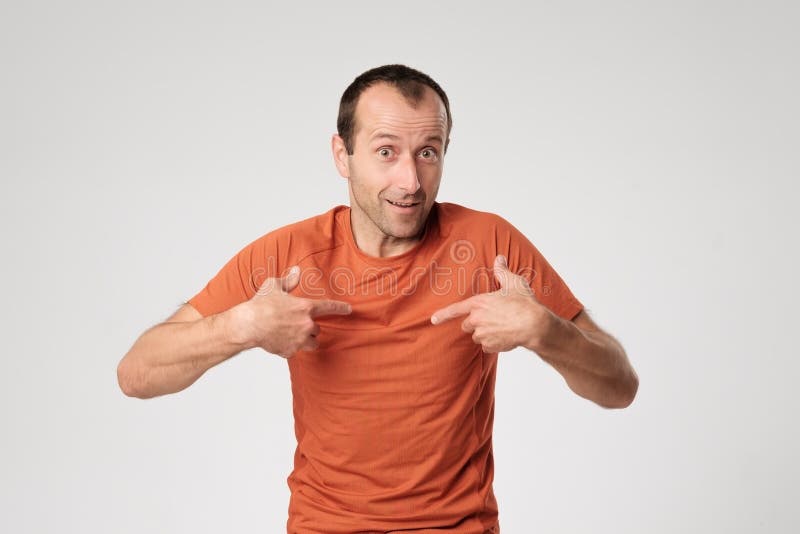 An adult Spanish man in an orange T-shirt points to himself with his finger.