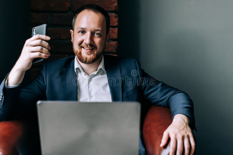Successful Businessman in suit sitting in a chair with a laptop, holding a smartphone in his hand and smiling stock image Successful Businessman in suit sitting in a chair with a laptop, holding a smartphone in his hand and smiling stock image