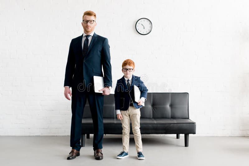 Adulto serio y hombres de negocios preadolescentes en las lentes que celebran los ordenadores portátiles y la mirada foto de archivo