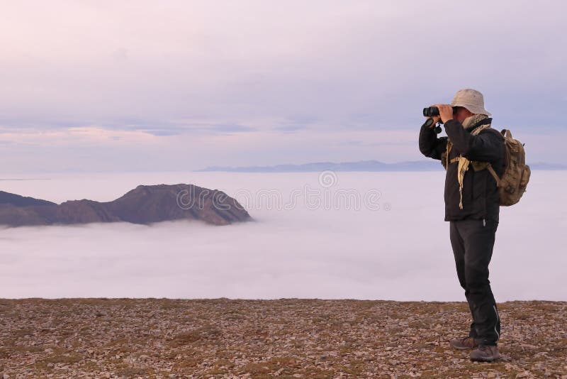 ADVENTURER MANN MIT HAT UND RÜCKHALTPACK AUSSIEHT DURCH BINOCULARS NEBEN EINEM TALLEY MIT SCHLAUBEN lizenzfreie stockfotos