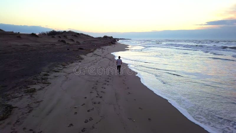 Aerial panning with drone on young attractive man jogging on the beach at sunrise. Running workout practice in fitness and healthy