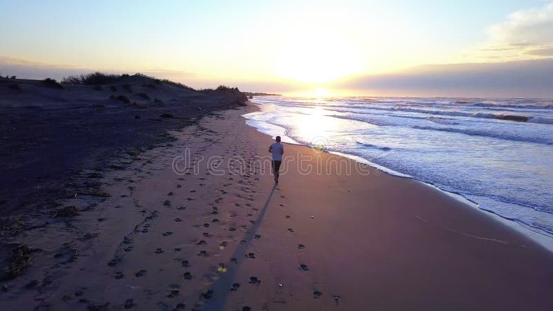 Aerial panning with drone on young attractive man jogging on the beach at sunrise. Running workout practice in fitness and healthy